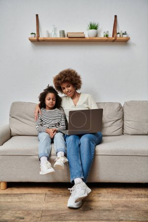 African American mother and daughter happily using a laptop while sitting on a couch at home.