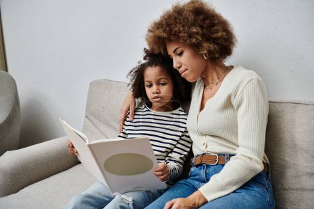 Photo for A happy African American mother and daughter sit on a couch, reading a book together, enjoying quality time at home. - Royalty Free Image