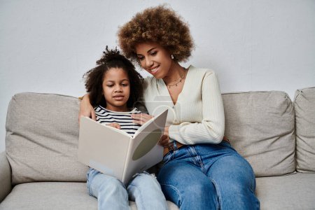 Una alegre madre e hija afroamericana inmersas en un libro cautivador mientras están sentadas cómodamente en un sofá.