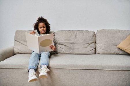 A little girl of African American descent sits on a cozy couch, engrossed in a book