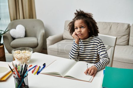 A young girl of African American descent sits at a table with a book and pens, immersed in her dreams