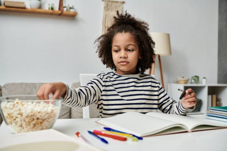 Foto de Una niña se sienta en una mesa con un tazón de palomitas de maíz y un libro, disfrutando de un momento de paz e imaginación - Imagen libre de derechos