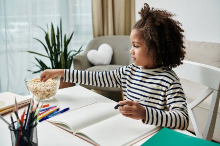 Téléchargez les photos : Une petite fille s'assoit joyeusement à une table, dégustant un bol de pop-corn - en image libre de droit