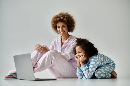 Téléchargez les photos : Une heureuse mère afro-américaine et sa fille en pyjama assis sur le sol à l'aide d'un ordinateur portable. - en image libre de droit