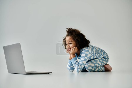 Téléchargez les photos : Une heureuse Afro-Américaine en pyjama est assise sur le sol, engloutie dans un ordinateur portable - en image libre de droit
