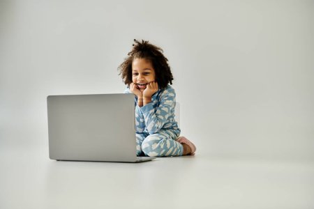 Ein junges afroamerikanisches Mädchen im Schlafanzug sitzt mit einem Laptop auf dem Boden und erkundet die digitale Welt.