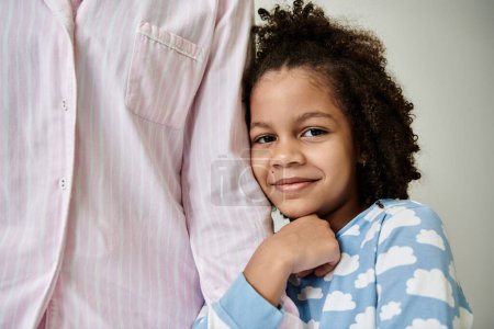 Una madre y una hija afroamericanas en pijamas acogedores posan felizmente juntas sobre un fondo gris.