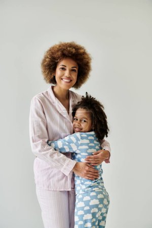 Happy African American mother and daughter sharing a warm hug in cozy pajamas on a grey background.