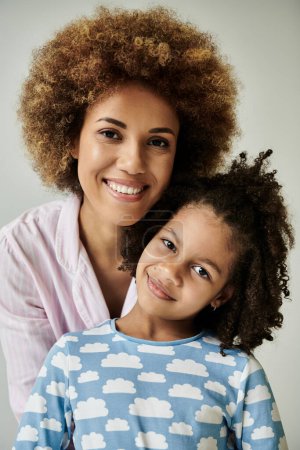 A happy African American mother and daughter pose together in matching pajamas against a grey background.