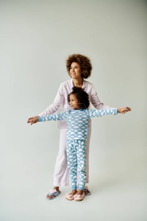 Fröhliche afroamerikanische Mutter und Tochter genießen die gemeinsame Zeit in kuscheligen Pyjamas vor grauem Hintergrund.