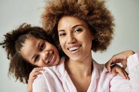 A joyful African American mother and daughter, dressed in pajamas, are striking a pose in front of a grey background.