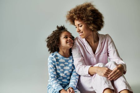 Eine glückliche afroamerikanische Mutter und Tochter im Pyjama sitzen zusammen auf einem grauen Hintergrund.