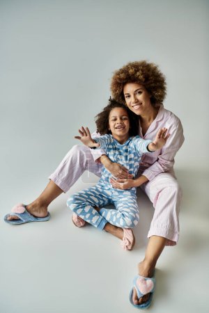 Foto de Una alegre madre afroamericana y su hija en pijama a juego posan juntas sobre un fondo gris. - Imagen libre de derechos