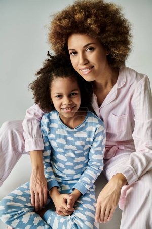 Foto de Sonriente madre e hija afroamericana posando en pijamas de colores sobre un suave fondo gris. - Imagen libre de derechos