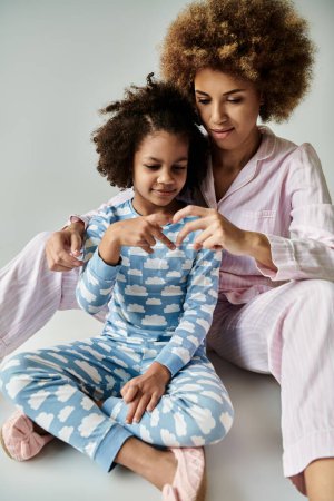 Joyful African American mother and daughter in matching pajamas sitting together on the floor.