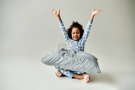 Joyful African American kid in pajamas, on a grey background. The little girl holds a pillow.