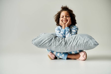Téléchargez les photos : Happy African American girl en pyjama, tenant un oreiller sur un fond gris. - en image libre de droit
