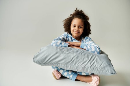 A joyful African American girl in pajamas holding a pillow, enjoying a cozy moment with her mother on a grey background.