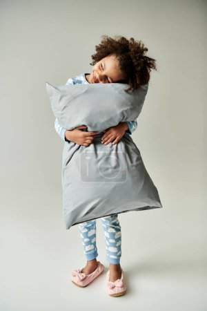 A young African American girl in pajamas hugs a pillow tightly in a peaceful moment against a gray backdrop.