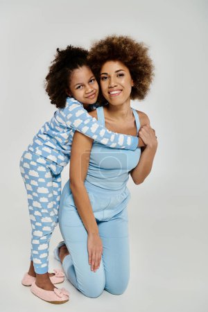 Embracing love and laughter, an African American mother and daughter strike a pose in matching blue pajamas on a grey backdrop.