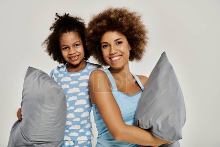 Téléchargez les photos : Bonne mère afro-américaine et sa fille en pyjama assorti tenant des oreillers sur un fond blanc. - en image libre de droit