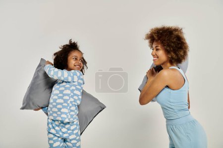 Foto de Feliz madre e hija afroamericana en pijama teniendo una juguetona pelea de almohadas sobre un fondo blanco. - Imagen libre de derechos