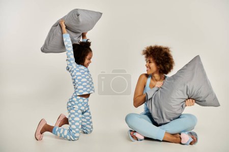 Foto de Una feliz madre afroamericana y su hija en pijama se involucran en una juguetona lucha de almohadas sobre un fondo gris. - Imagen libre de derechos