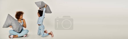 Joyful African American mother and daughter in pajamas playfully tossing pillows in a cozy moment on a grey background.