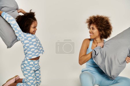 Madre e hija afroamericanas en pijama jugando a lanzar almohadas en el aire sobre un fondo blanco.