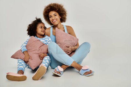 Téléchargez les photos : Bonne mère afro-américaine et sa fille assis sur le sol en pyjama, profiter d'un moment confortable ensemble sur un fond gris. - en image libre de droit