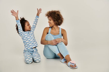 An ecstatic African American mother and daughter in pajamas sitting on the floor, arms raised to the sky in unison.