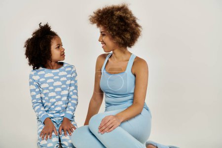 Foto de Una feliz madre afroamericana y su hija en pijama azul sentadas sobre un fondo blanco. - Imagen libre de derechos