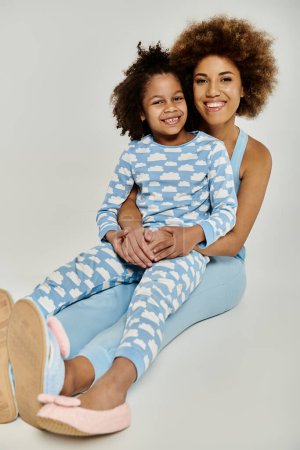Happy African American mother and daughter sitting on the floor in matching blue pajamas, sharing a cozy moment together.