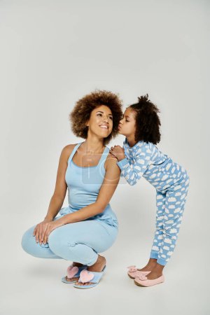 Téléchargez les photos : Un moment affectueux entre une joyeuse mère afro-américaine et sa fille, portant un pyjama bleu assorti, partageant un baiser. - en image libre de droit