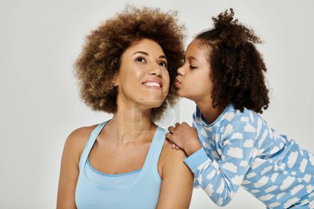 Happy African American mother and daughter wearing pajamas share a tender kiss in front of a white background.