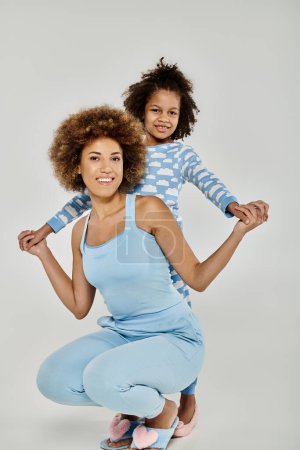 Sonriente madre e hija afroamericana usando pijamas azules a juego posan juntas sobre un fondo gris.