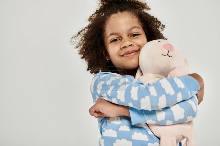 Foto de African American girl in pajamas cuddling a large stuffed animal on a grey background. - Imagen libre de derechos