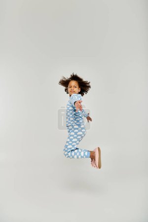 An energetic young girl, dressed in blue pajamas, leaps joyfully in the air on a grey background.