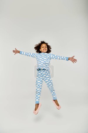 Una chica afroamericana feliz saltando alegremente en un pijama azul con su madre cerca sobre un fondo gris.