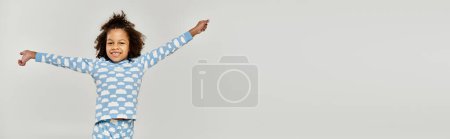 Una alegre chica afroamericana en pijama extendiendo sus brazos sobre un fondo gris.