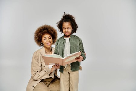 An African American mother reads a book to her little daughter, surrounded by love and joy in stylish clothing on a grey background.