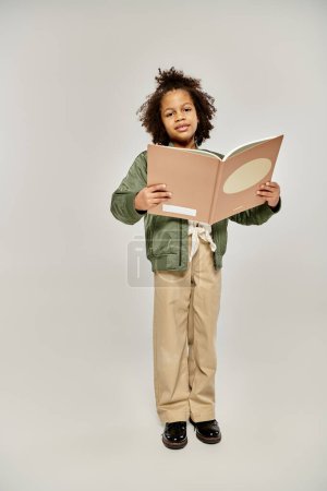 A young girl with a book, captivated by the pages, standing against a white backdrop.