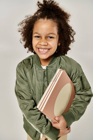Joven chica afroamericana en una chaqueta verde bombardero sosteniendo un libro.