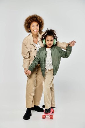 Téléchargez les photos : Élégant frisé afro-américain mère et fille posant gracieusement sur une planche à roulettes sur un fond gris. - en image libre de droit