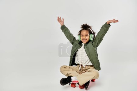 Photo for A curly African American girl in stylish clothes sits on a skateboard with her arms up in excitement. - Royalty Free Image