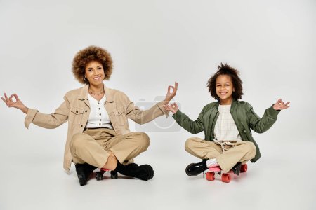 Foto de Madre e hija afroamericanas rizadas en trajes elegantes, sentadas en monopatines, practicando yoga juntas sobre un fondo gris. - Imagen libre de derechos
