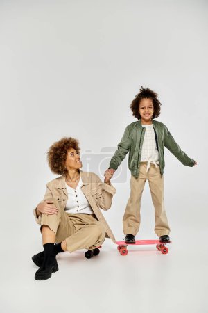 Curly African American mother and daughter in stylish clothes enjoy a fun skateboarding session together on a grey background.