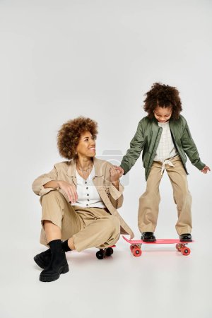 Photo for A curly African American mother and daughter in stylish clothes enjoy skateboarding together on a grey background. - Royalty Free Image
