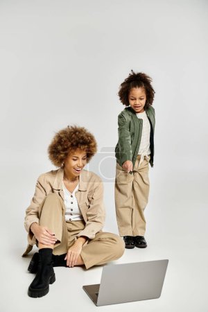 Téléchargez les photos : Curly African American mother and daughter sit on floor, deeply focused on laptop screen in stylish attire on grey background. - en image libre de droit