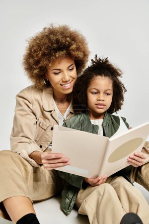 Eine lockige afroamerikanische Mutter und Tochter in stilvoller Kleidung genießen einen gemütlichen Moment beim gemeinsamen Lesen eines Buches auf grauem Hintergrund.